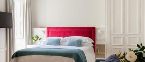 Habitación de hotel con cabecera roja, cama con ropa de cama blanca y azul, mesita de noche con lámpara, televisor montado en la pared y ventana con cortinas.