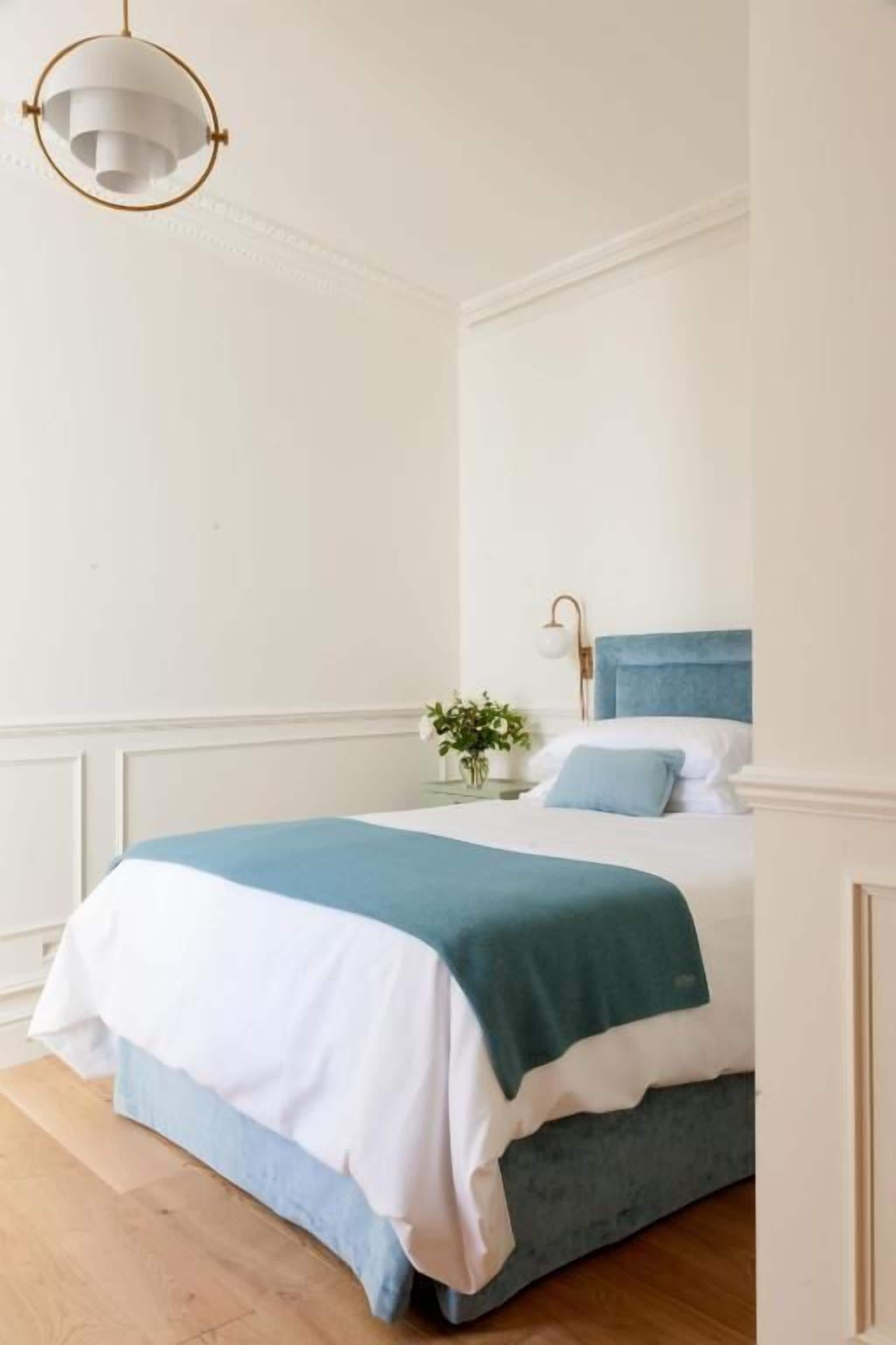 Una cama bien tendida con edredón azul y blanco, una mesita de noche con lámpara, y un colgante de techo.