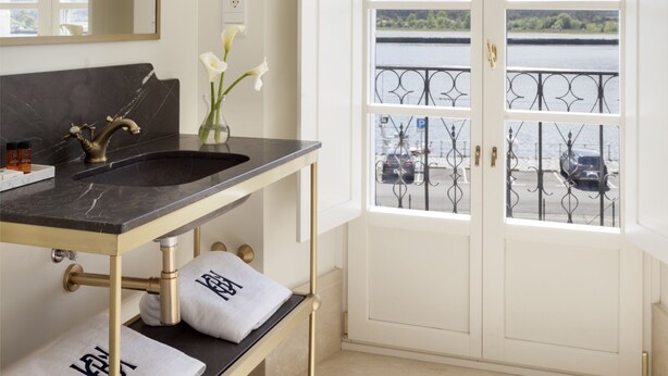 A bathroom with a black marble sink, gold fixtures, and a view of a lake through a white door with a balcony. - Gran Hotel Brillante (Muros de Nalon)