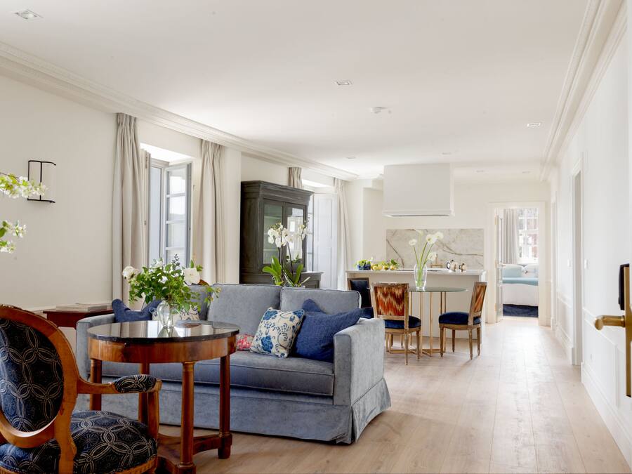Een ruime woonkamer met een grijze bank, houten stoelen, een ronde salontafel, en een eethoek met een witte tafel en stoelen.