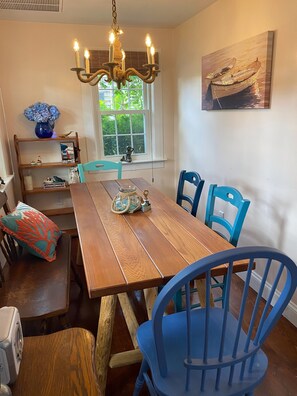 A dining area with a wooden table, blue chairs, a chandelier, a window with a view, and a bookshelf with decorative items. - Walk to the Beach! (Dennis Port)