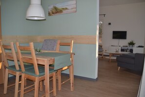 Ein Essbereich mit Holztisch und -stühlen, einer grünen Tischdecke und einer weißen Pendelleuchte. An der Wand ist ein Fernseher angebracht und auf einem Beistelltisch steht eine Pflanze.