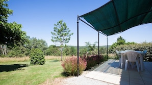Un patio al aire libre con una mesa y sillas bajo un toldo verde, rodeado de césped y árboles.