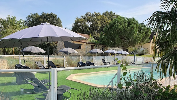 Zona junto a la piscina con sillones de descanso y sombrillas, rodeada de vegetación y un edificio de piedra.