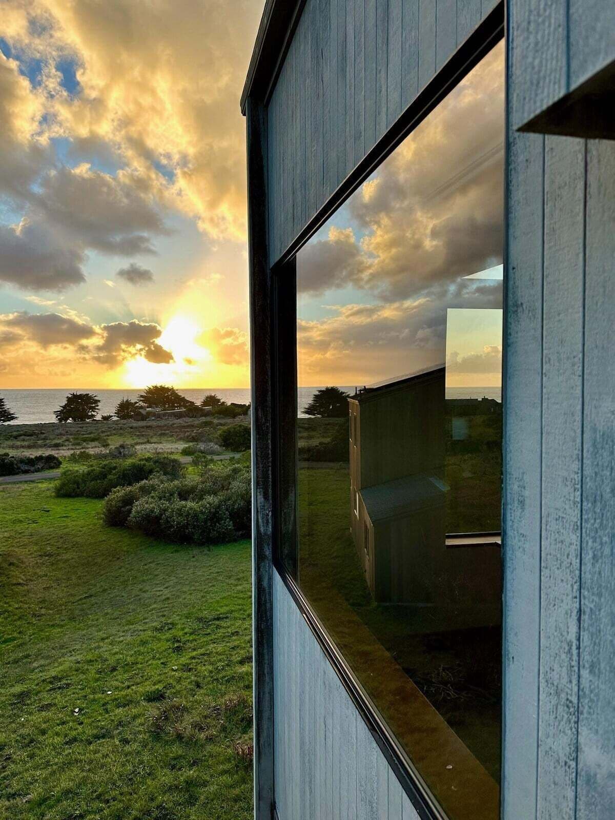 En moderne bygning med et stort vindue, der spejler en solnedgang over et græslandskab.
