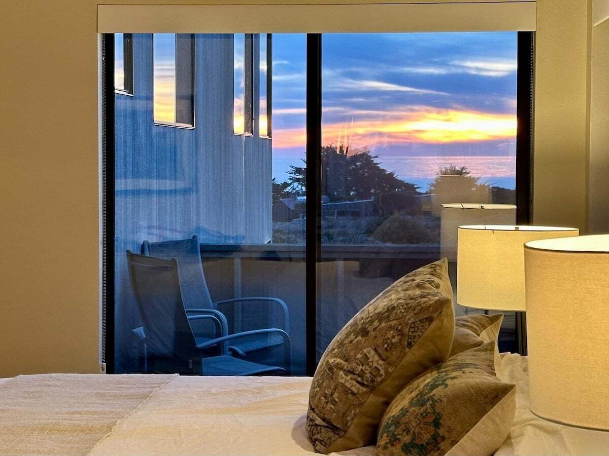 A hotel room with a bed, a window with a view of a building and trees, and a chair outside.