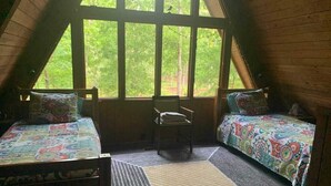 Une chambre mansardée chaleureuse, avec deux lits, un fauteuil et une grande fenêtre donnant sur une forêt.