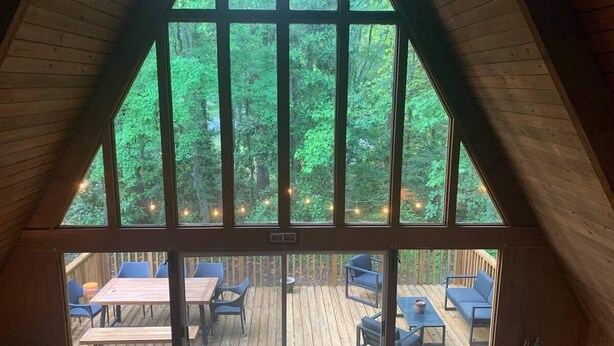 Une terrasse en bois avec des sièges extérieurs et une vue sur une forêt, visible à travers de grandes fenêtres.