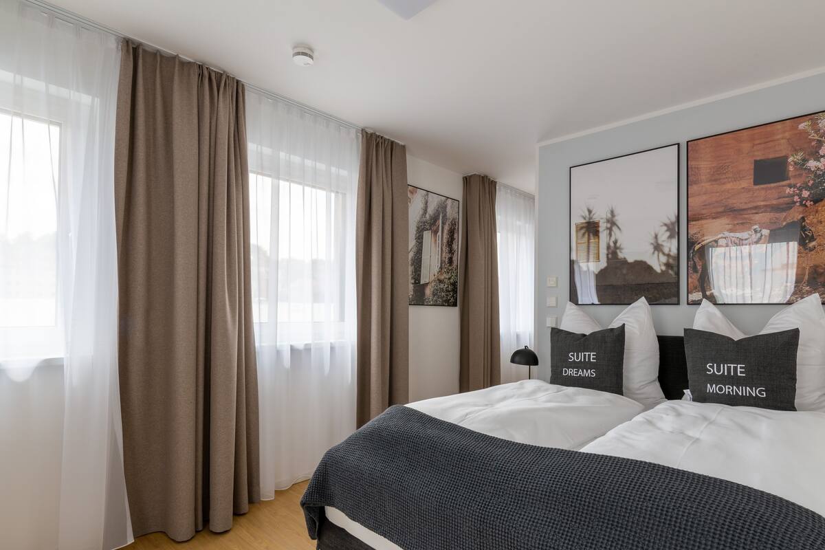 Moderni hotellihuone, jossa on suuri sänky, kaksi koristetyynyä, joissa lukee ”SUITE DREAMS” ja ”SUITE MORNING”, musta lattiavalaisin ja taideteoksia seinillä.