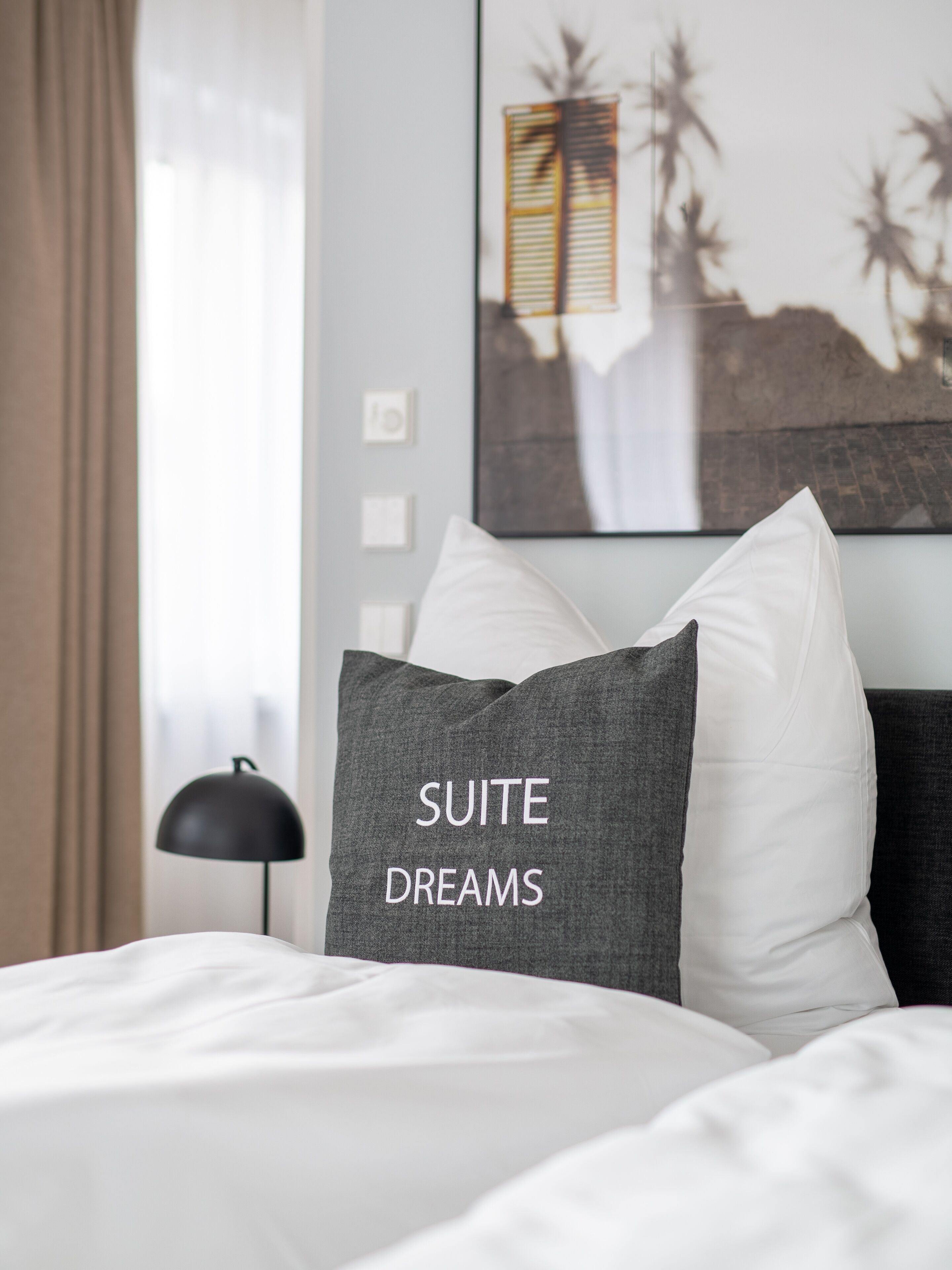 A hotel room with a bed, a pillow with "SUITE DREAMS" embroidered, a lamp, and a wall with a framed picture.