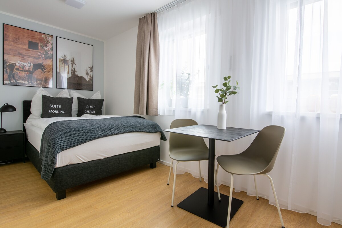 A modern hotel room with a bed, bedside tables, a desk with chairs, and large windows with sheer curtains.