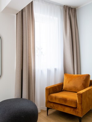 A room with a sofa and a round ottoman, a window with sheer curtains, and a wall-mounted mirror. - Limehome Passau Bahnhofstraße (Passau)