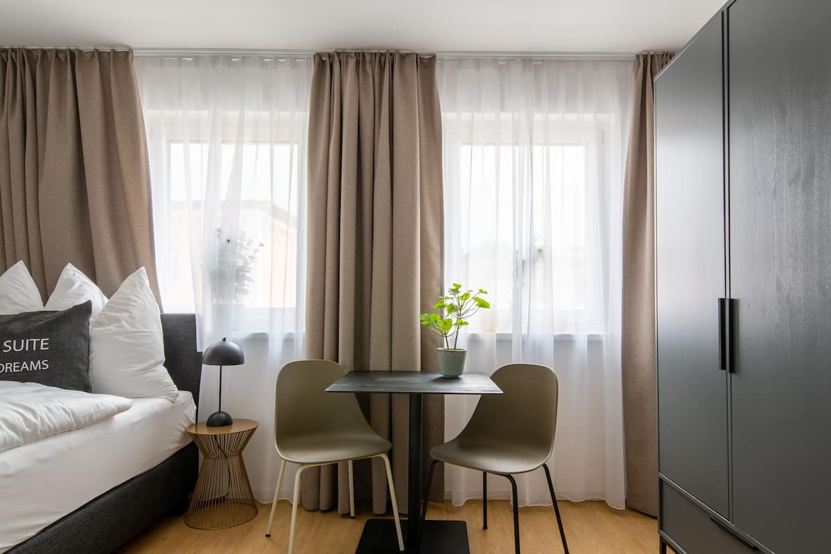 Moderni hotellihuone, jossa on sänky, pieni pöytä tuoleineen, ruukkukasvi ja vaatekaappi.