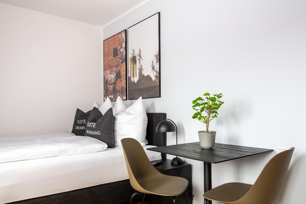 Moderni hotellihuone, jossa on sänky, kaksi tuolia, pieni pöytä, ruukkukasvi ja taideteos seinällä.