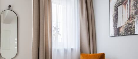 A room with a large window, a chair, a round ottoman, and a wall-mounted mirror.