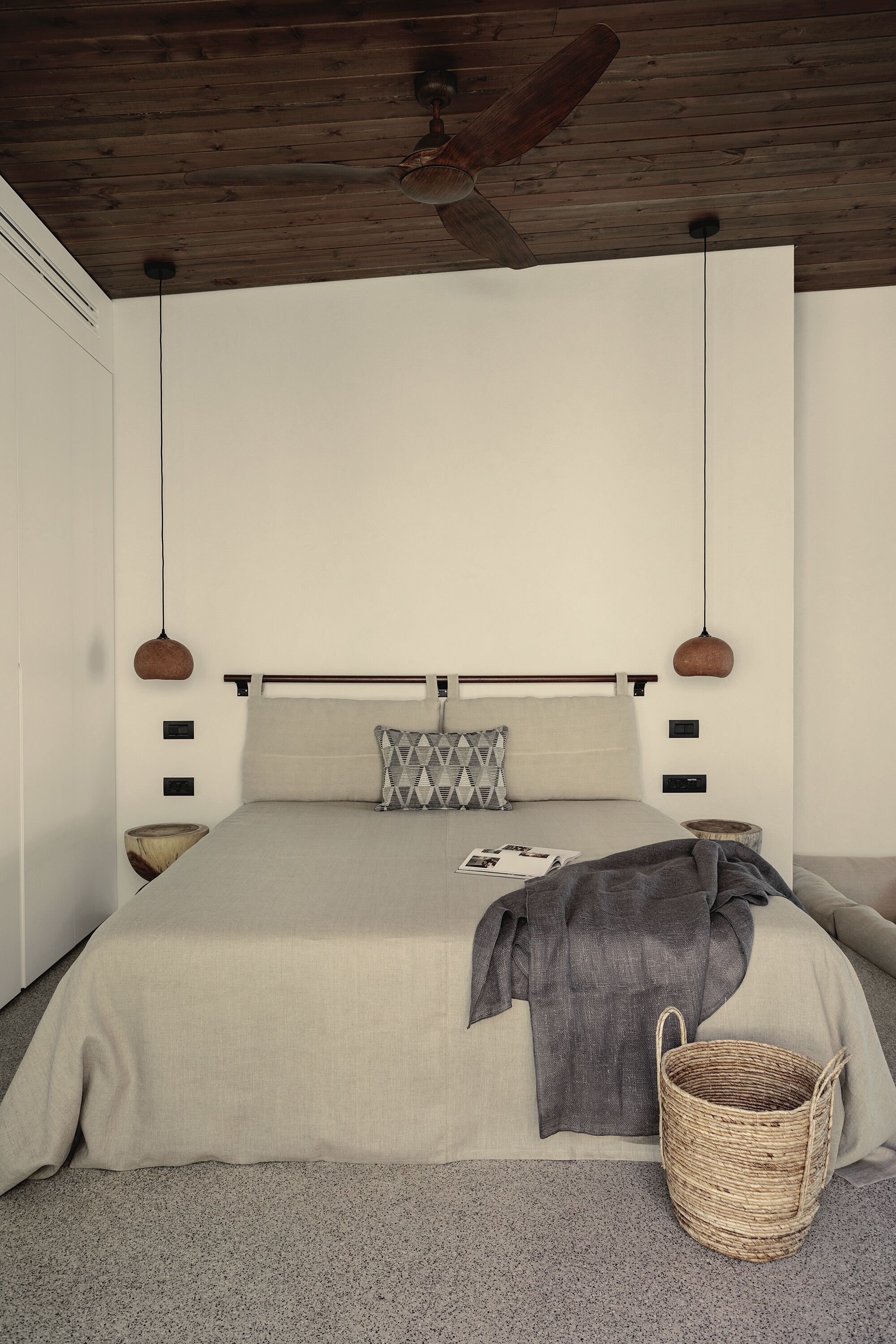 Un dormitorio con techo de madera, una cama con manta gris, almohada gris y una cesta tejida en el suelo.