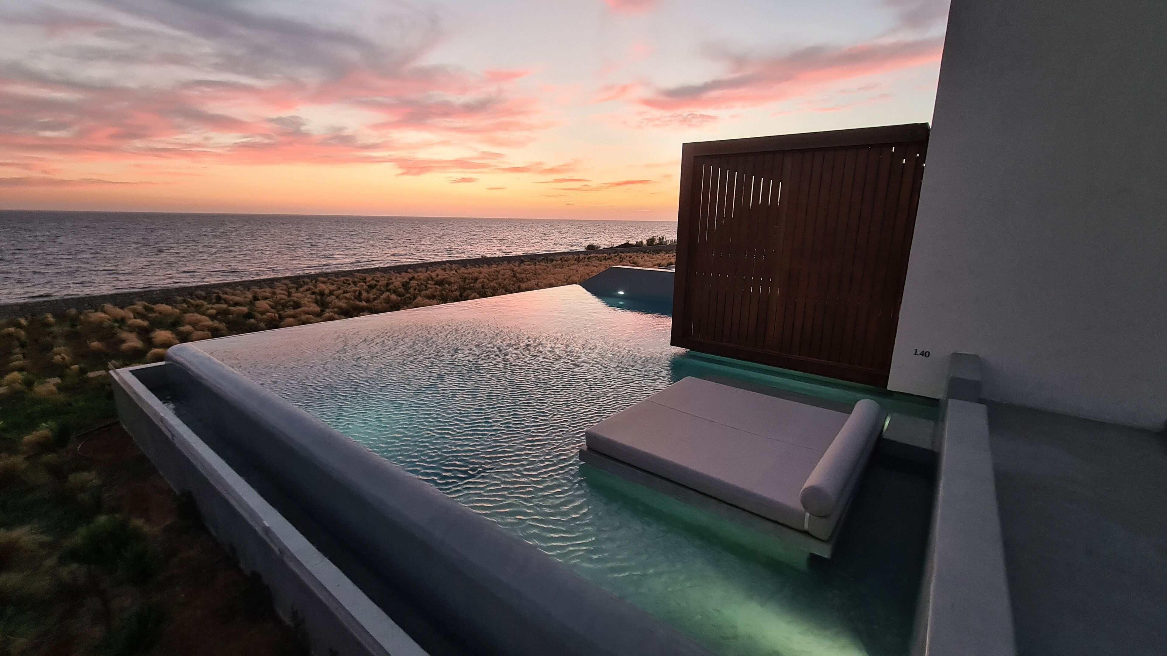 A infinity pool with a view of the ocean at sunset, a wooden deck, and a lounge area with a cushion.