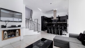 Een moderne woonkamer met een open haard, een flatscreen televisie, een grijze bank en een keuken met zwarte apparaten en barkrukken.