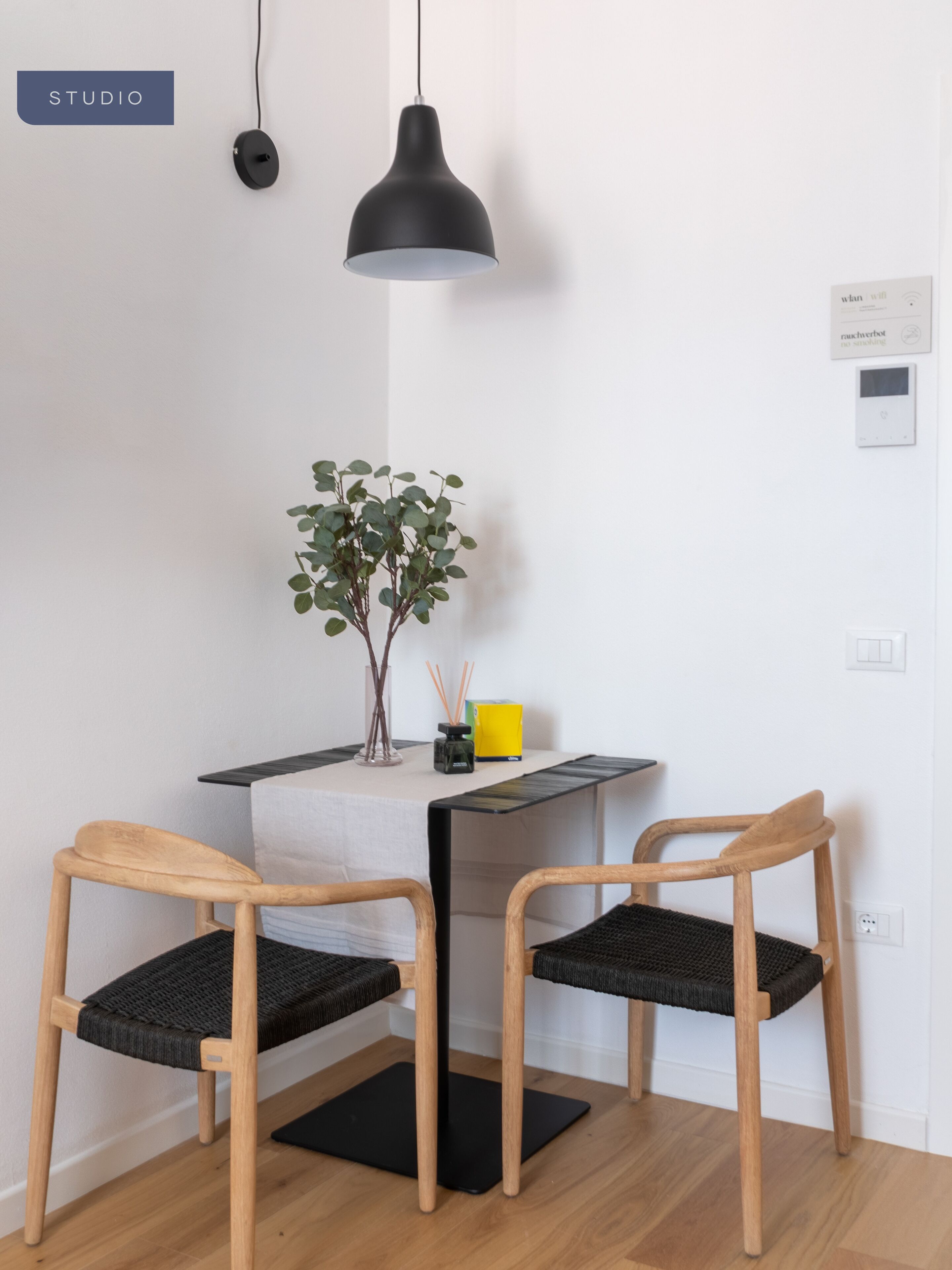 Een minimalistisch ingerichte kamer met twee houten stoelen, een klein tafeltje, een plant in pot en een zwart hanglampje.