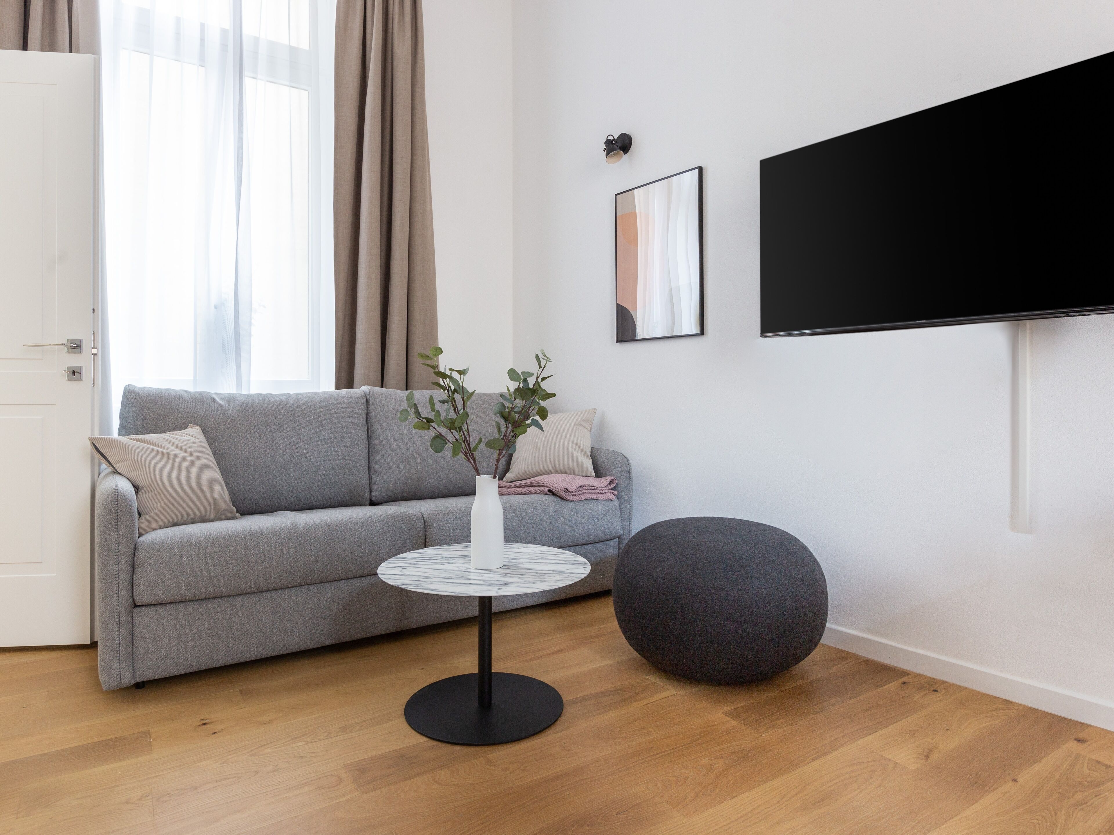 Una sala de estar moderna con un sofá gris, una mesa de centro redonda, un otoman gris, un jarrón blanco con plantas, un cuadro enmarcado y un televisor grande en la pared.
