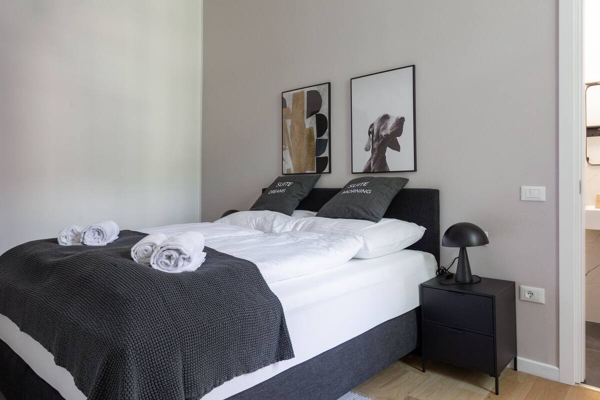 Una habitación de hotel moderna con una cama bien tendida, una mesita de noche con lámpara y dos cuadros enmarcados en la pared.