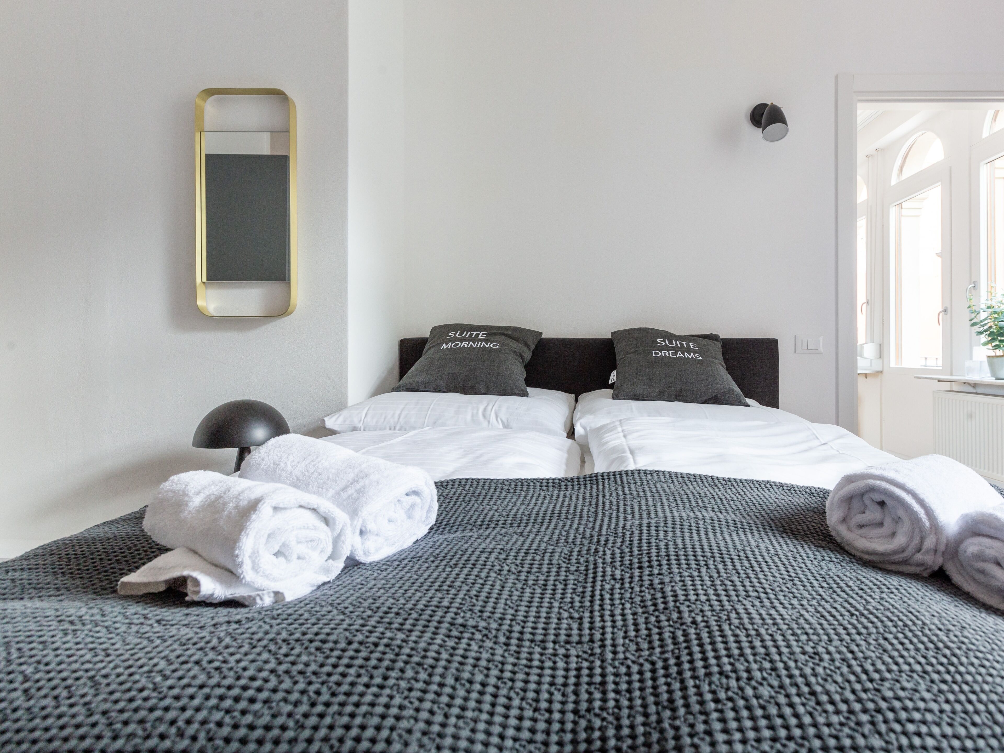 Cama arrumada com duas almofadas, manta cinza texturizada e toalhas brancas enroladas.
