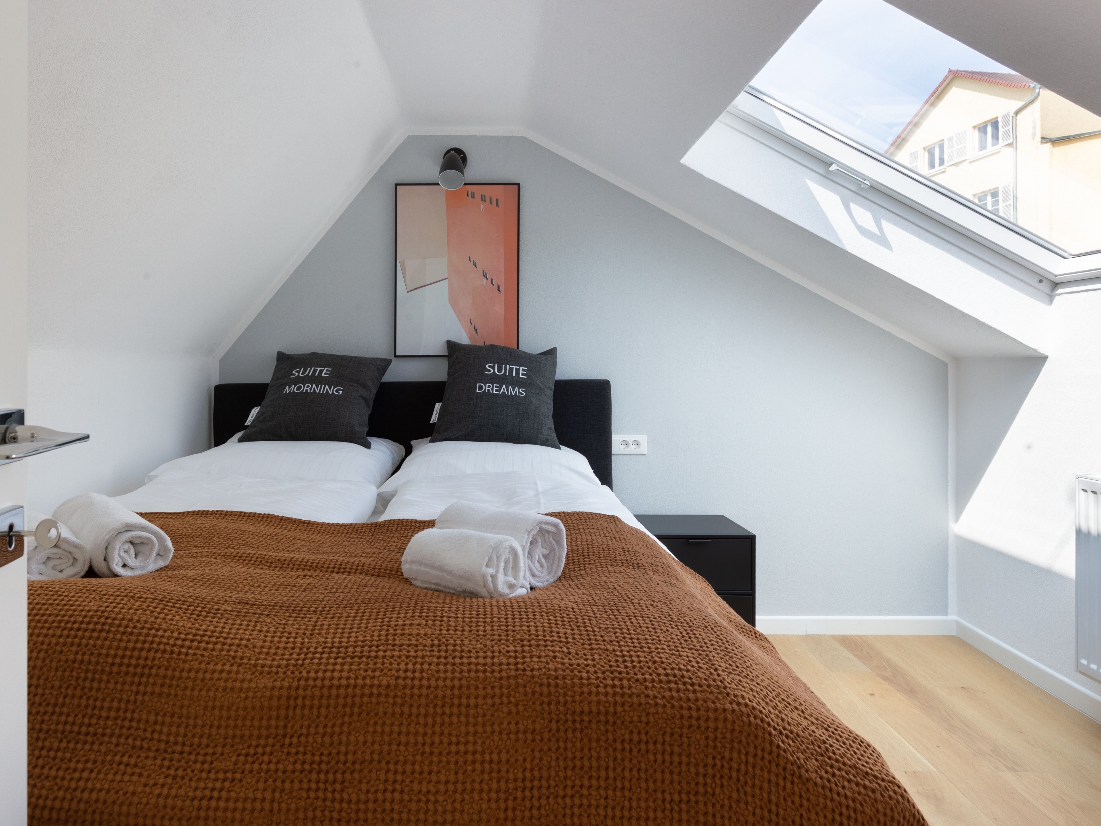 Un dormitorio moderno con tragaluces, dos camas con almohadas a juego, una manta marrón y una obra de arte enmarcada en la pared.