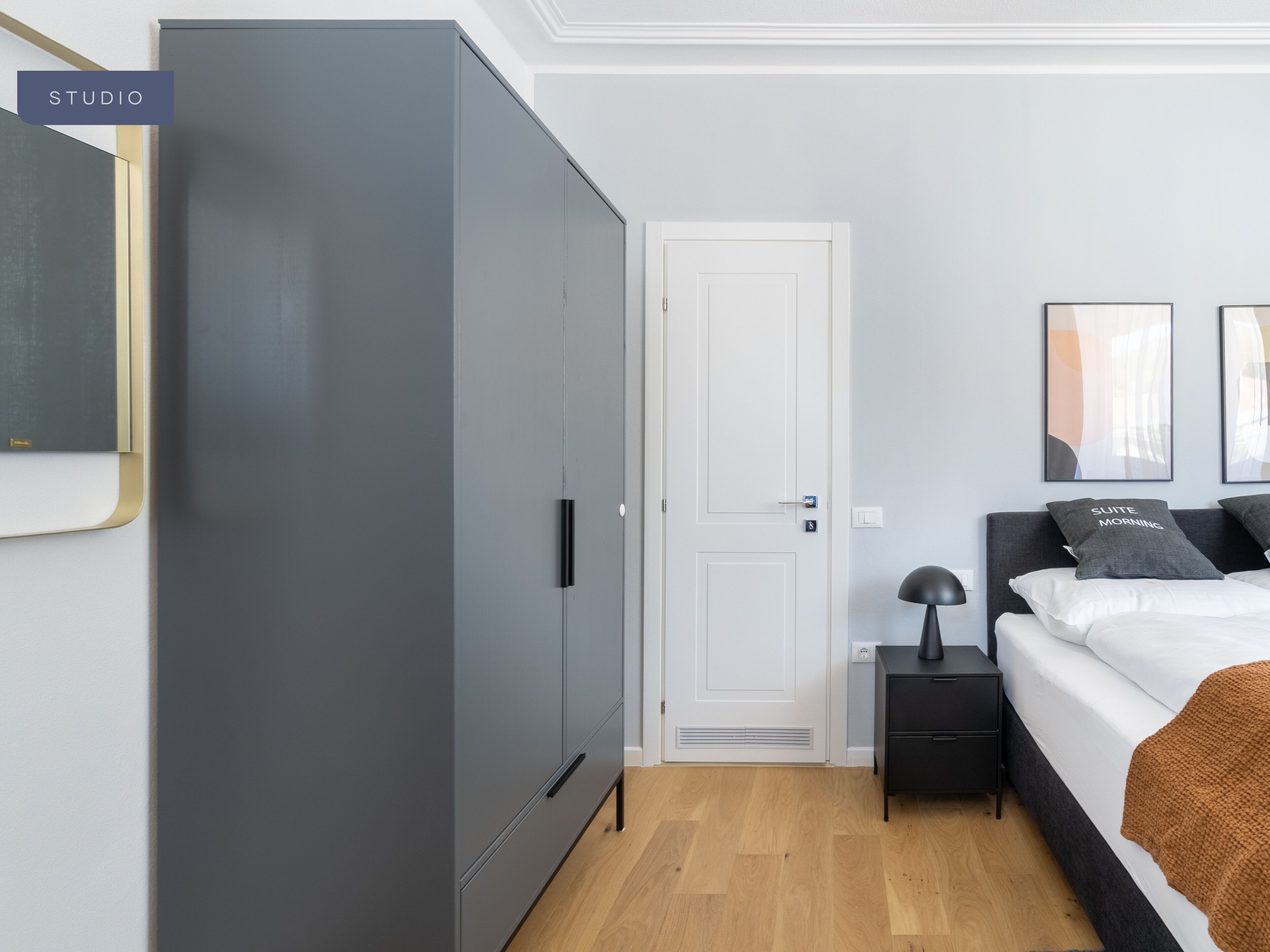 Un dormitorio con un armario gris, una puerta blanca, una cama con cabecero negro, una mesita de noche y dos cuadros en la pared.