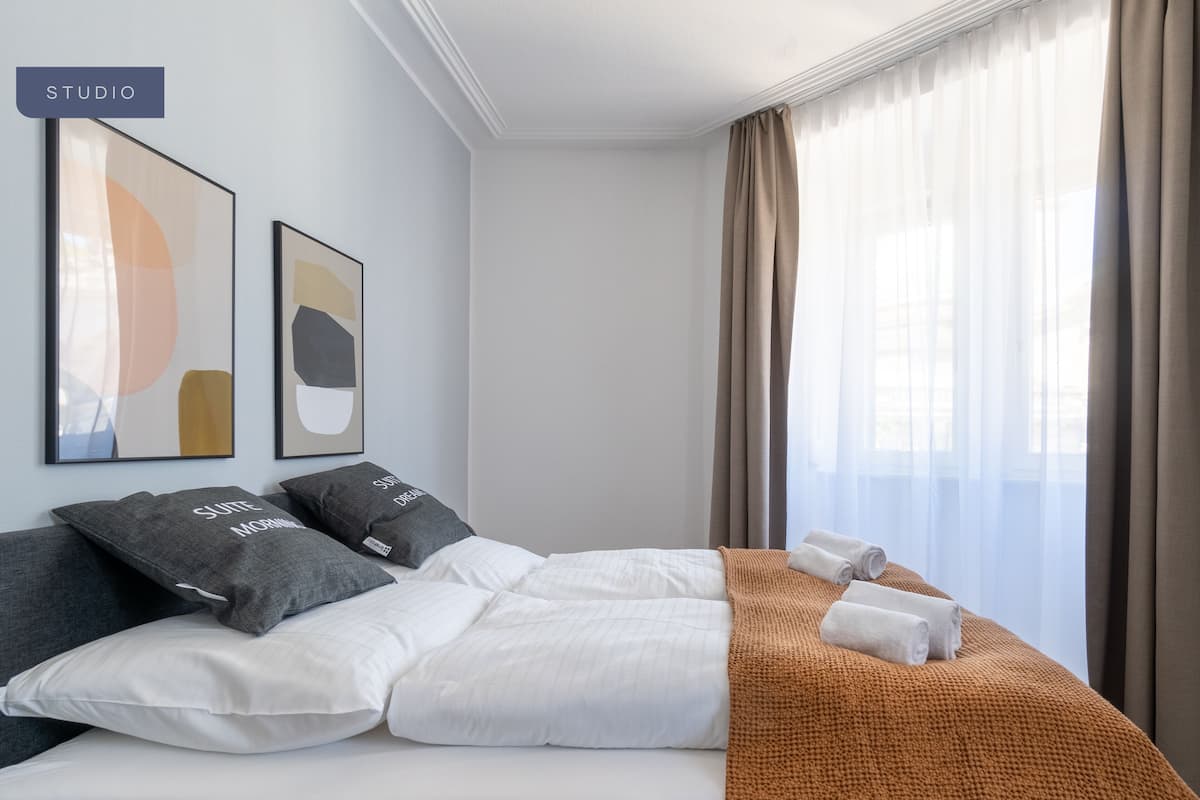 Habitación de hotel con una cama, dos almohadas, una colcha, una ventana con cortinas y dos obras de arte abstractas en la pared.