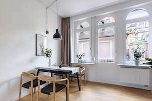 Uma área de refeições com mesa, cadeiras e uma luminária pendente. Há janelas com cortinas e vista para edifícios.