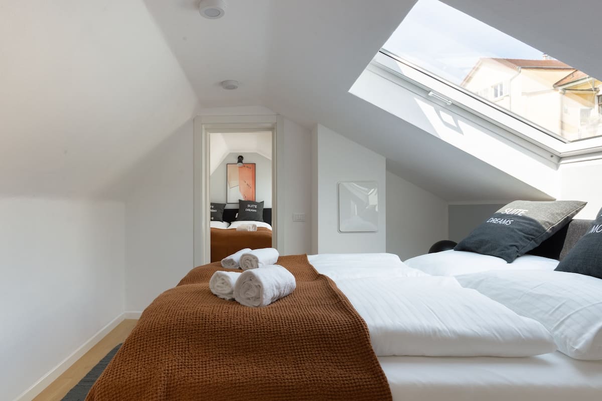 Habitación de hotel con tragaluz, cama con sábanas blancas, manta marrón y vistas a la ciudad desde la ventana.