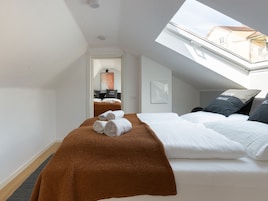 Quarto de hotel com claraboia, cama com lençóis brancos, manta marrom e vista da cidade pela janela.