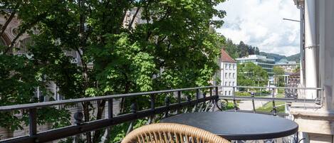 Un balcón con una silla de mimbre y una mesa, con vista a un paisaje urbano con edificios y árboles.