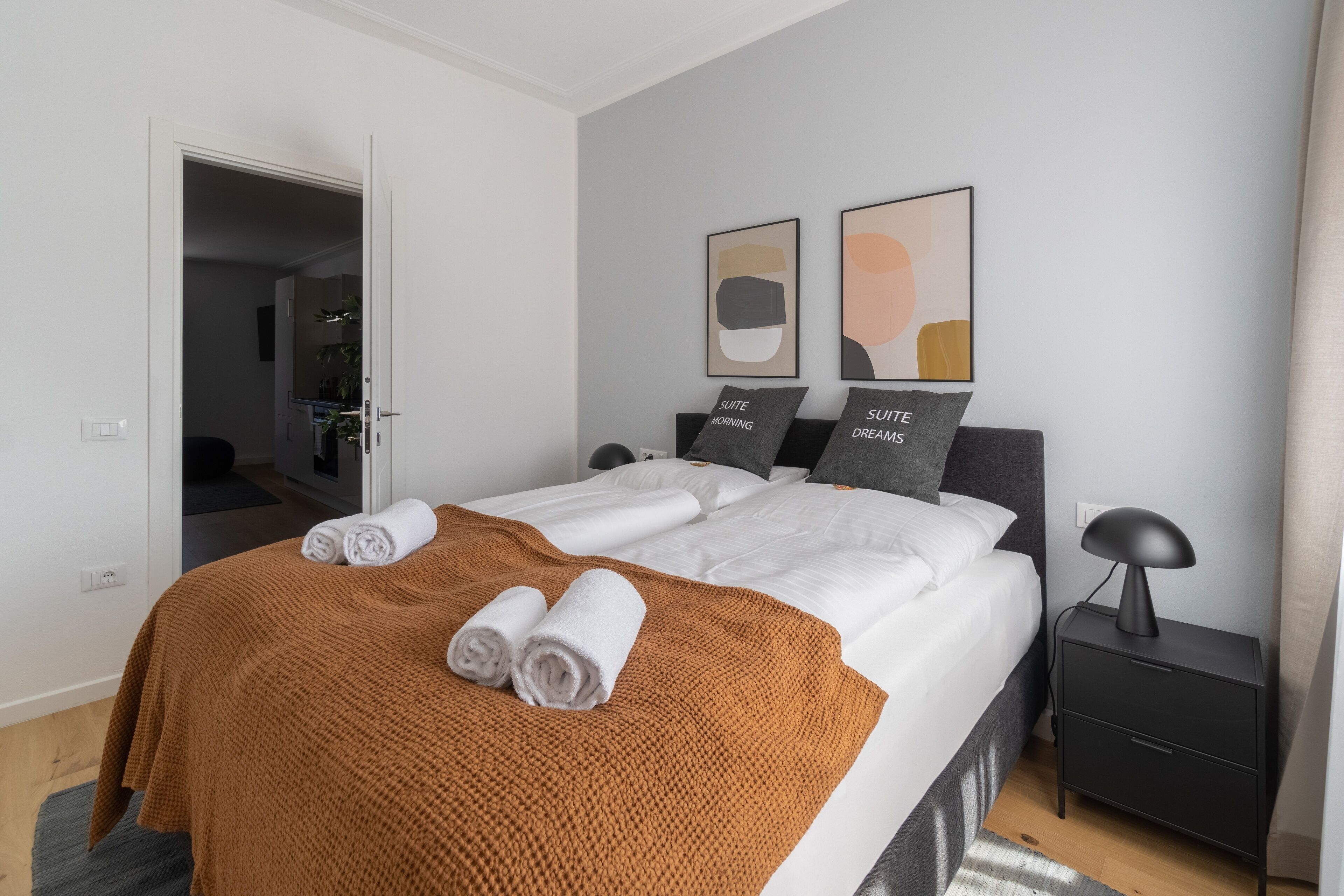 A modern hotel room with a neatly made bed, a brown throw blanket, and two pillows with text. There are two framed abstract paintings on the wall.