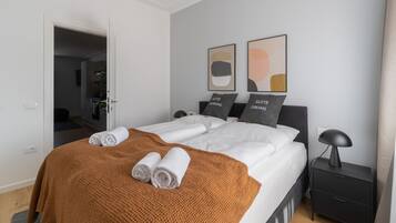 A modern hotel room with a neatly made bed, a brown throw blanket, and two pillows with text. There are two framed abstract paintings on the wall.