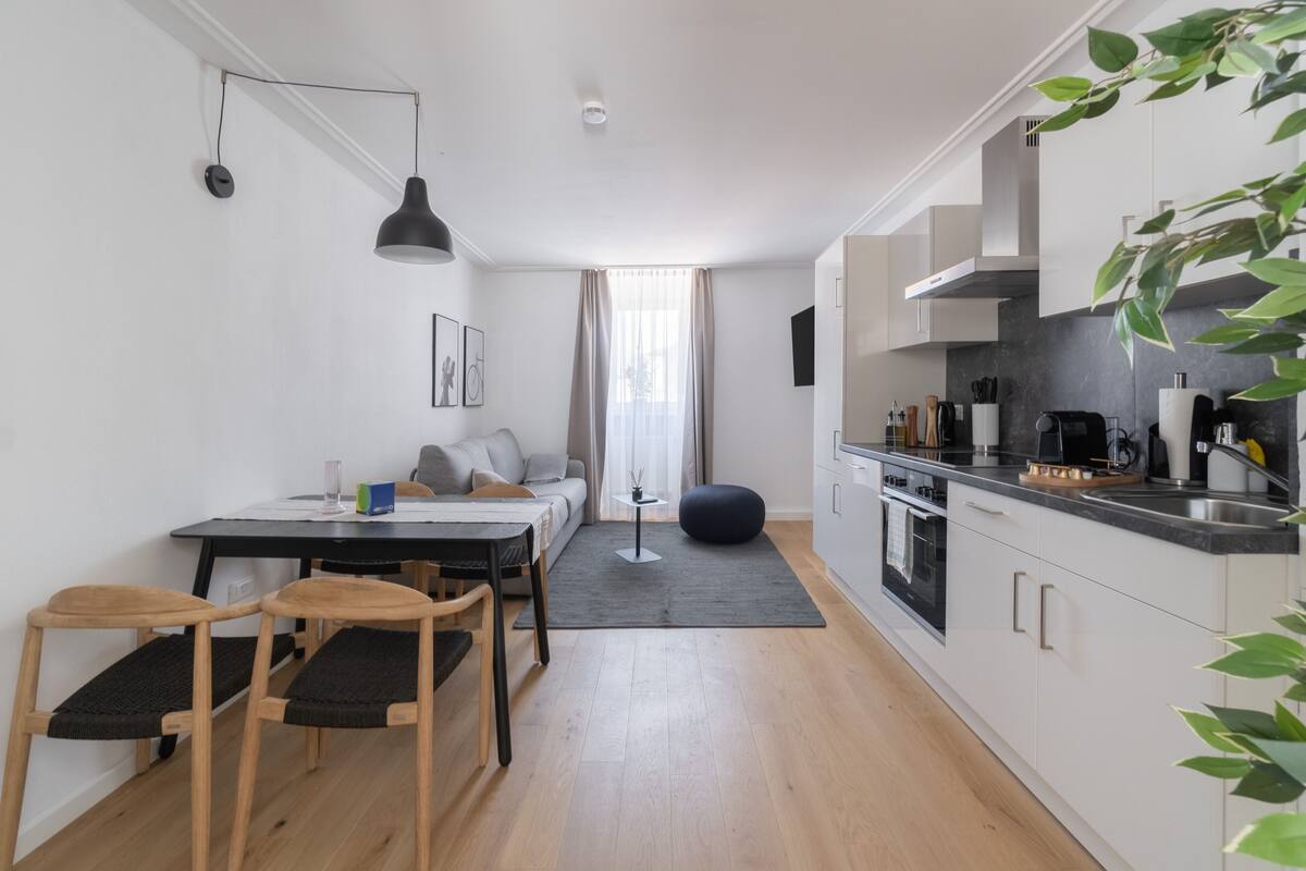 Una cocina moderna con armarios blancos, encimera negra y zona de comedor con mesa y sillas.