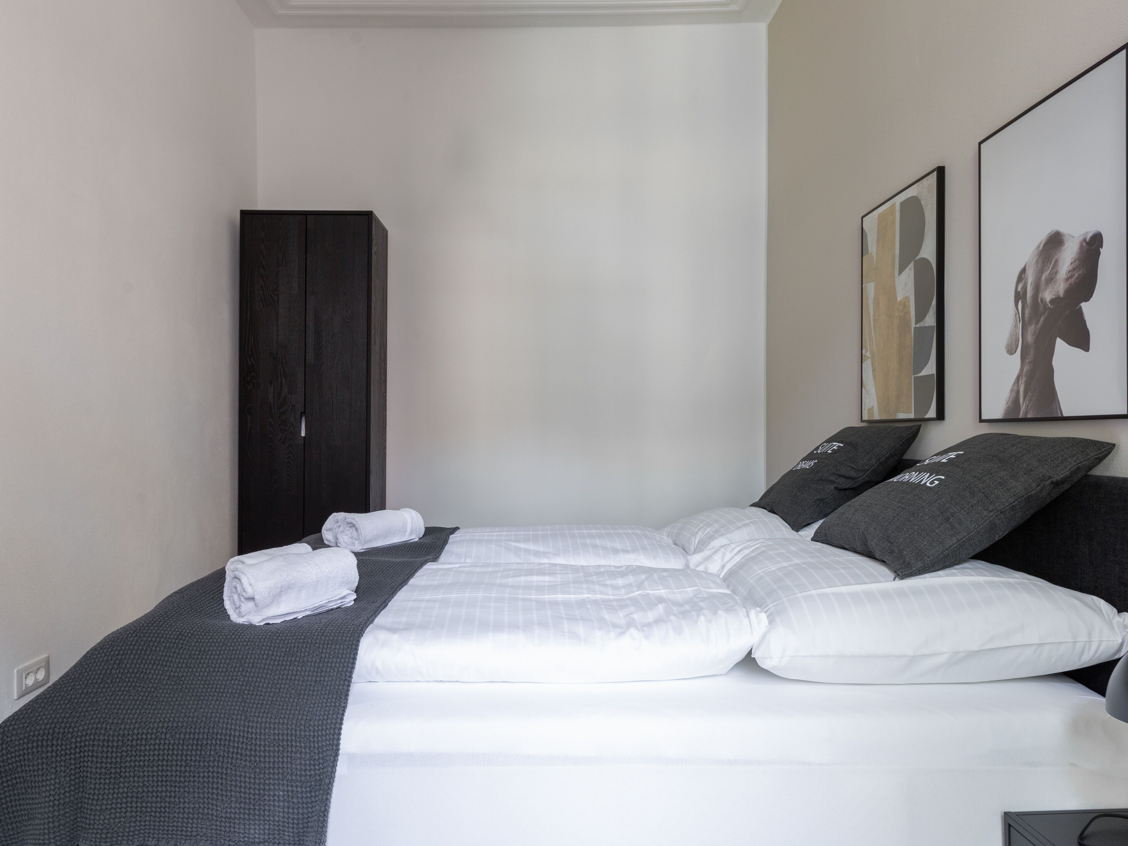 Una cama bien hecha con sábanas blancas y almohadas grises, un armario de madera oscura y dos cuadros enmarcados en la pared.