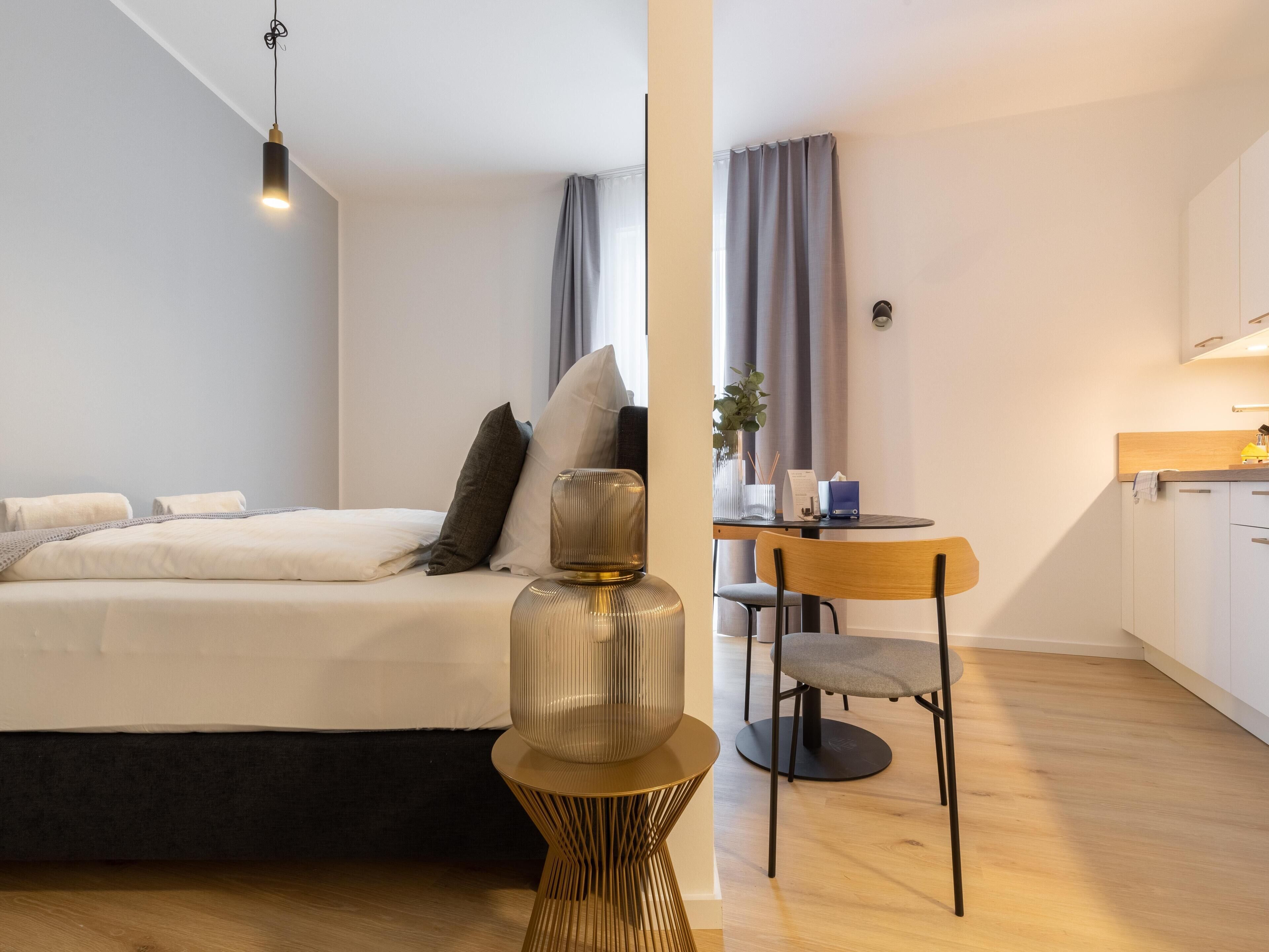 A modern bedroom with a bed, bedside table, and a small dining area with a chair and table.
