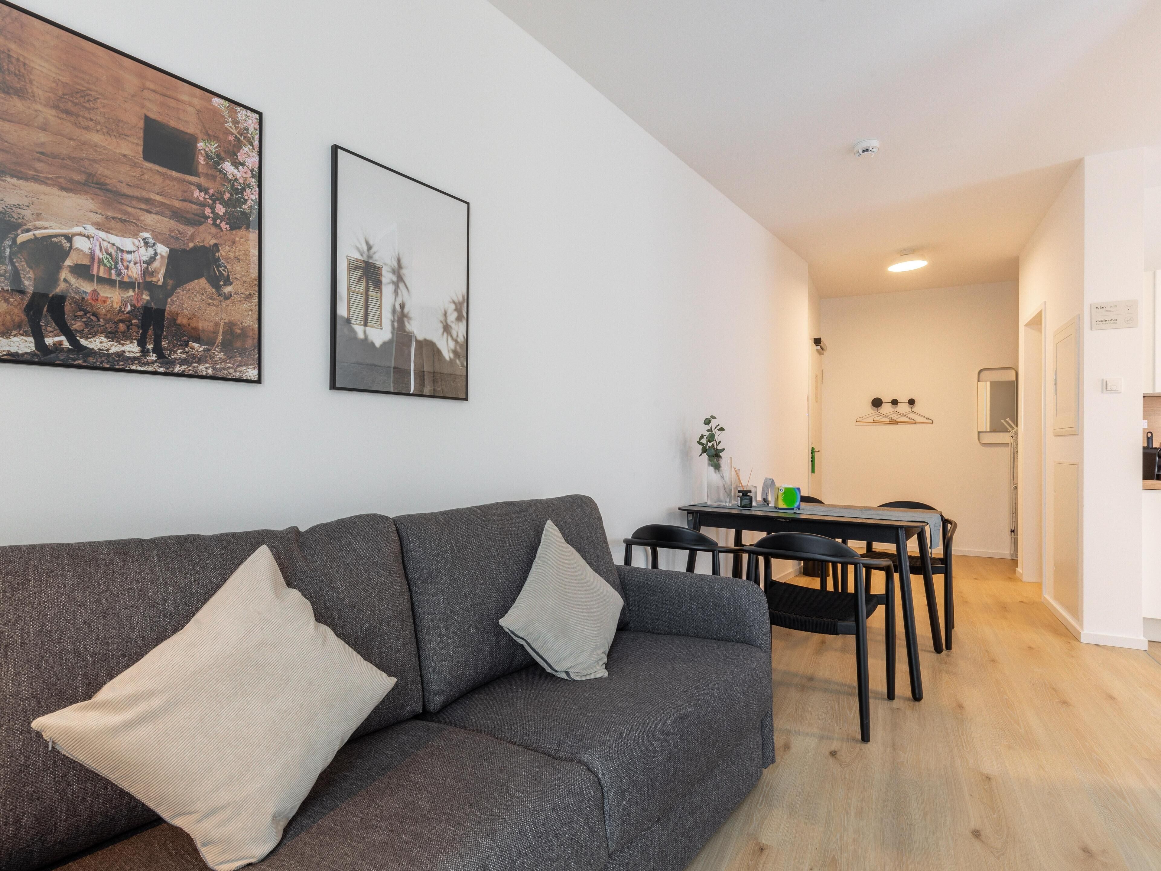 Una sala de estar moderna con un sofá gris, dos cojines, una mesa de comedor con sillas y una cocina al fondo.