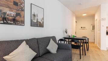 Una sala de estar moderna con un sofá gris, dos cojines, una mesa de comedor con sillas y una cocina al fondo.