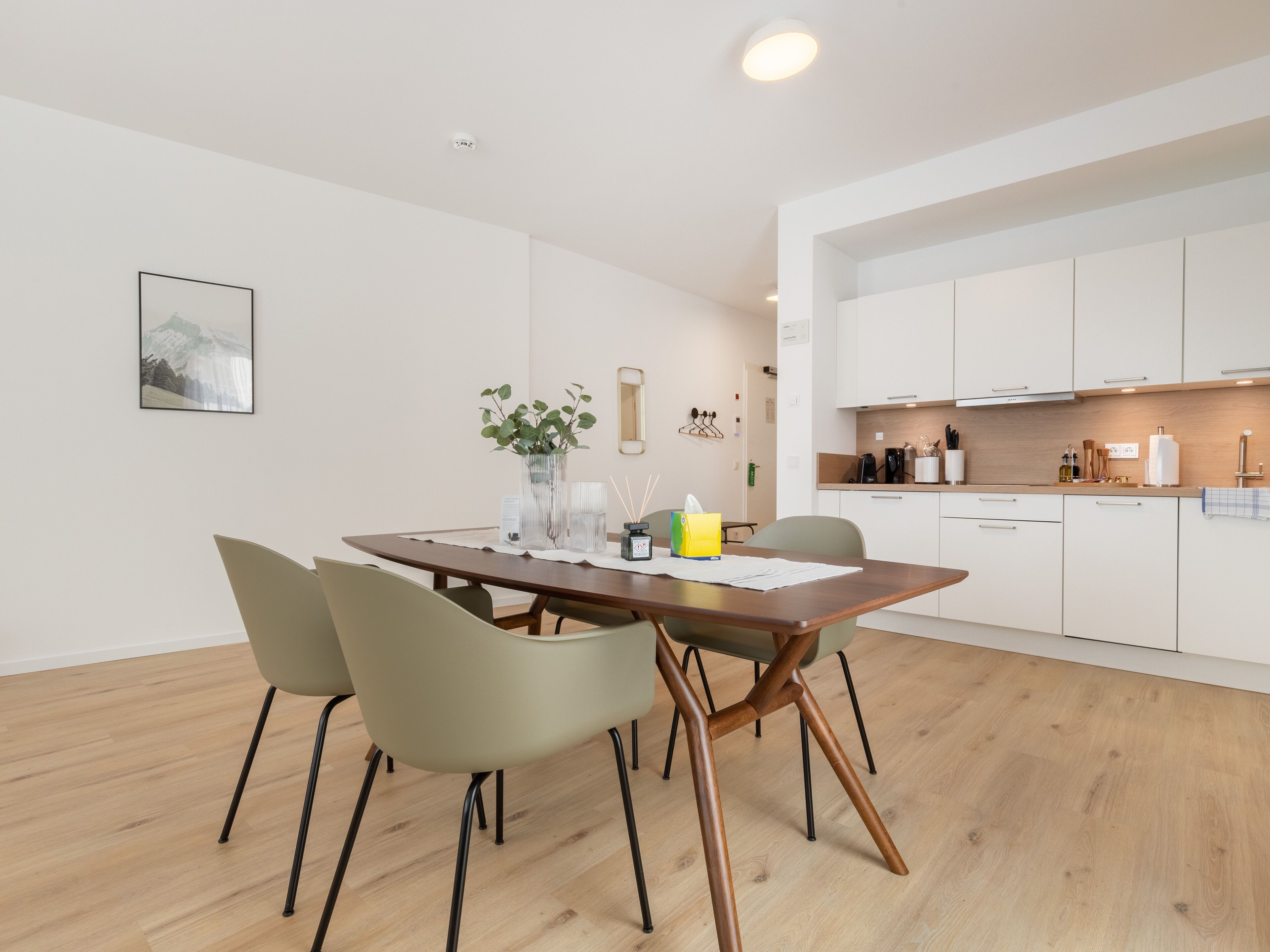 Un área de comedor moderna con una mesa de madera, cuatro sillas, un florero con plantas y un cuadro en la pared.