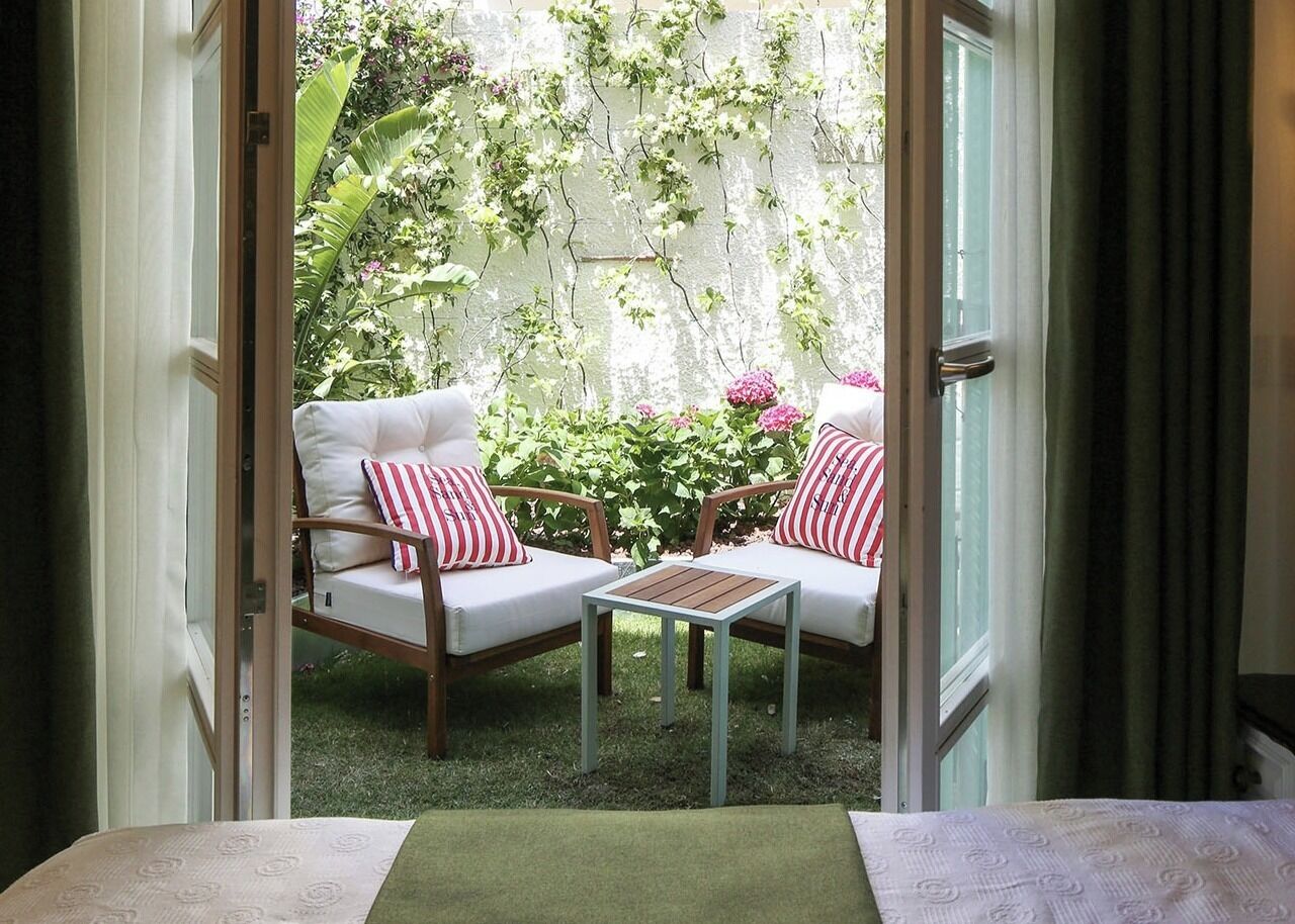 A room with two white cushioned chairs with red and white striped pillows, a small wooden table, and a view of a lush garden through an open door.