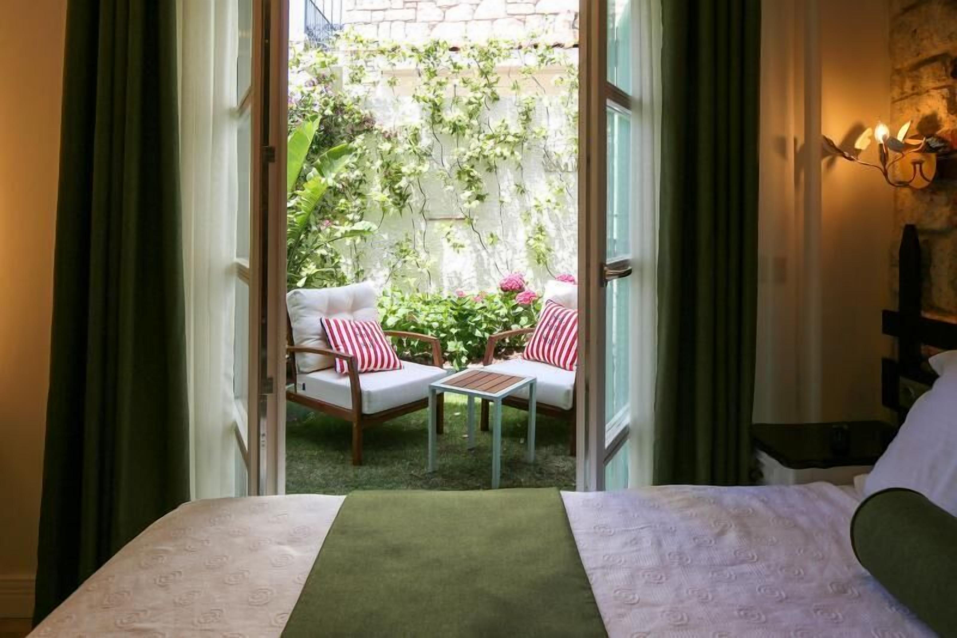 Ein Hotelzimmer mit Bett, einem kleinen Tisch, zwei Sesseln mit rot-weiß gestreiften Kissen und Blick auf einen Garten durch eine offene Tür.