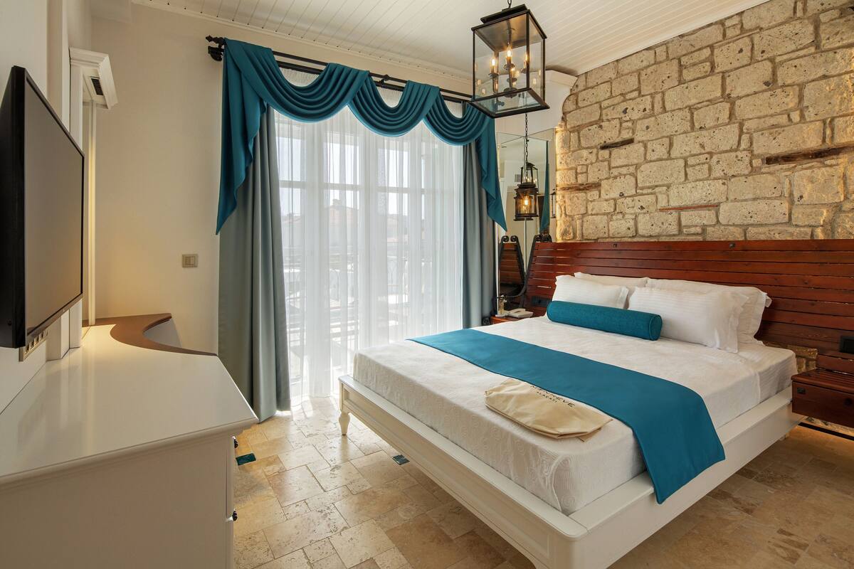 A hotel room with a large bed, a television, a stone wall, and a window with curtains.