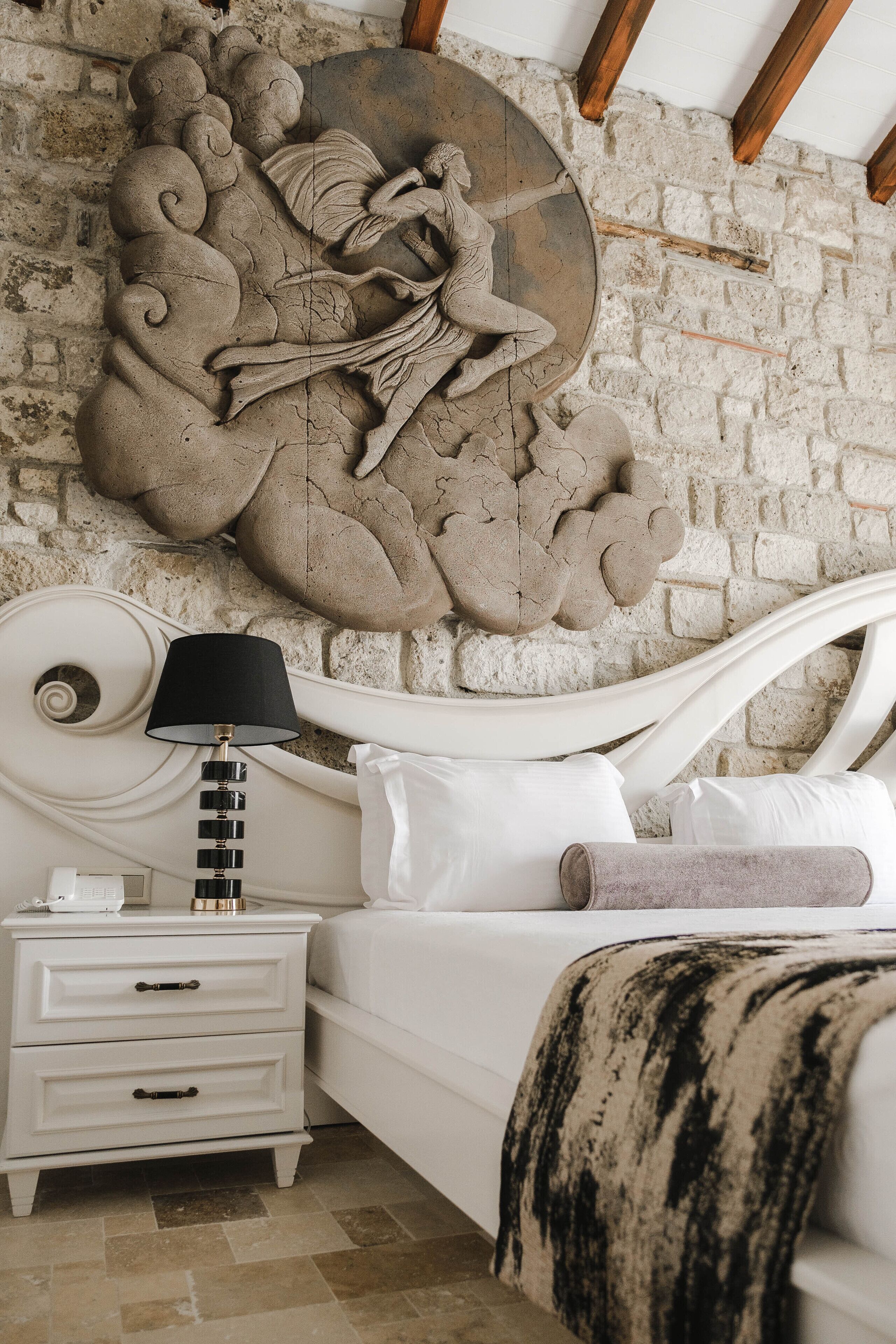 A bedroom with a white bed, a nightstand with a lamp, and a stone wall sculpture.