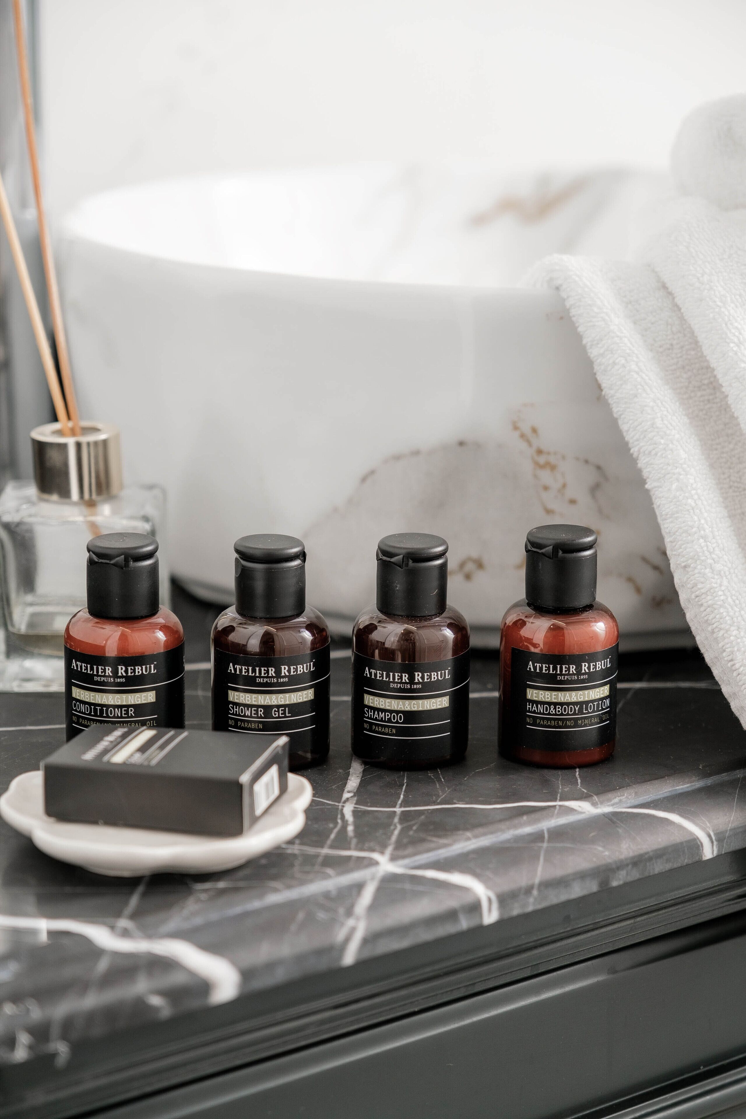 Four bottles of Atelier Rebul products on a marble surface with a white basin and a white towel in the background.