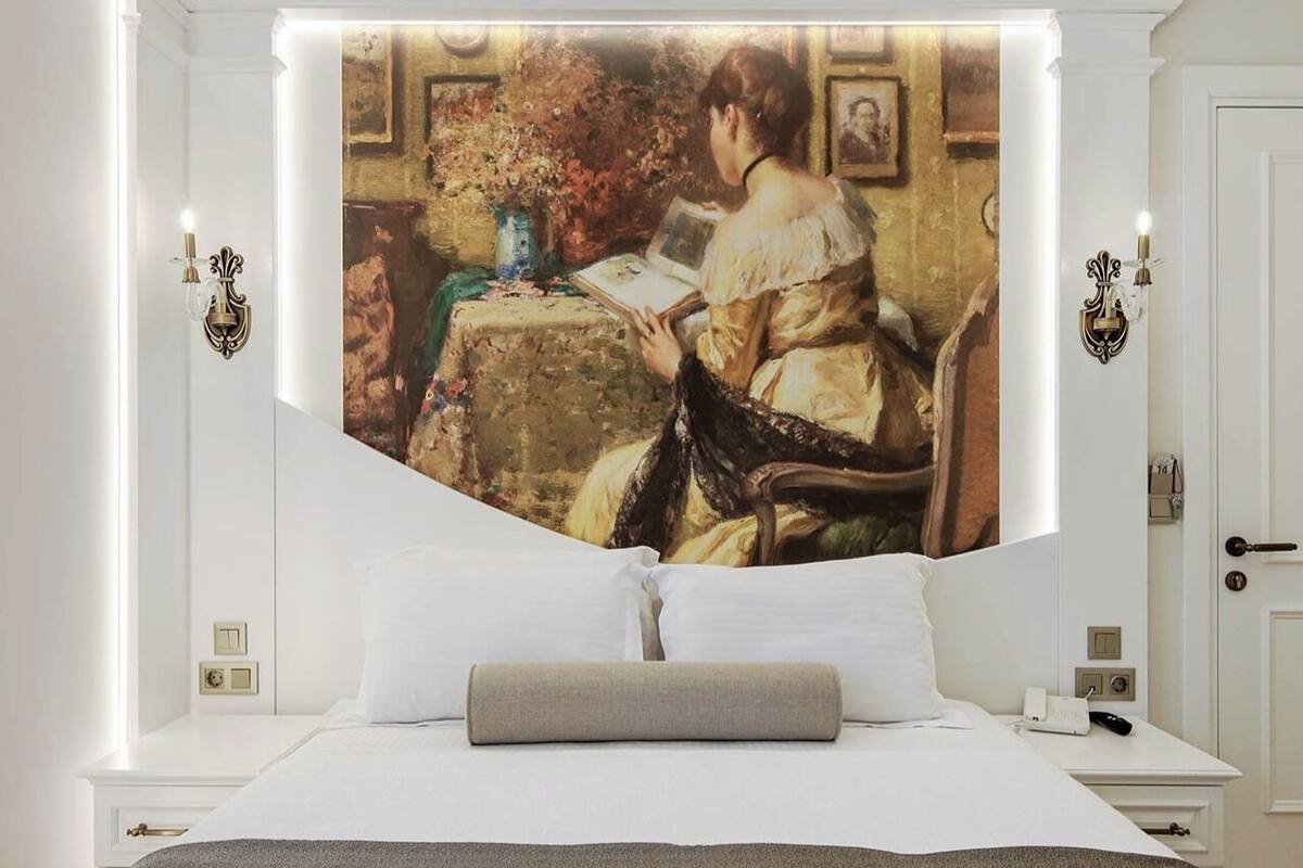 A bedroom with a large painting of a woman reading, white walls, a bed with white linens, and a bedside table with a lamp.