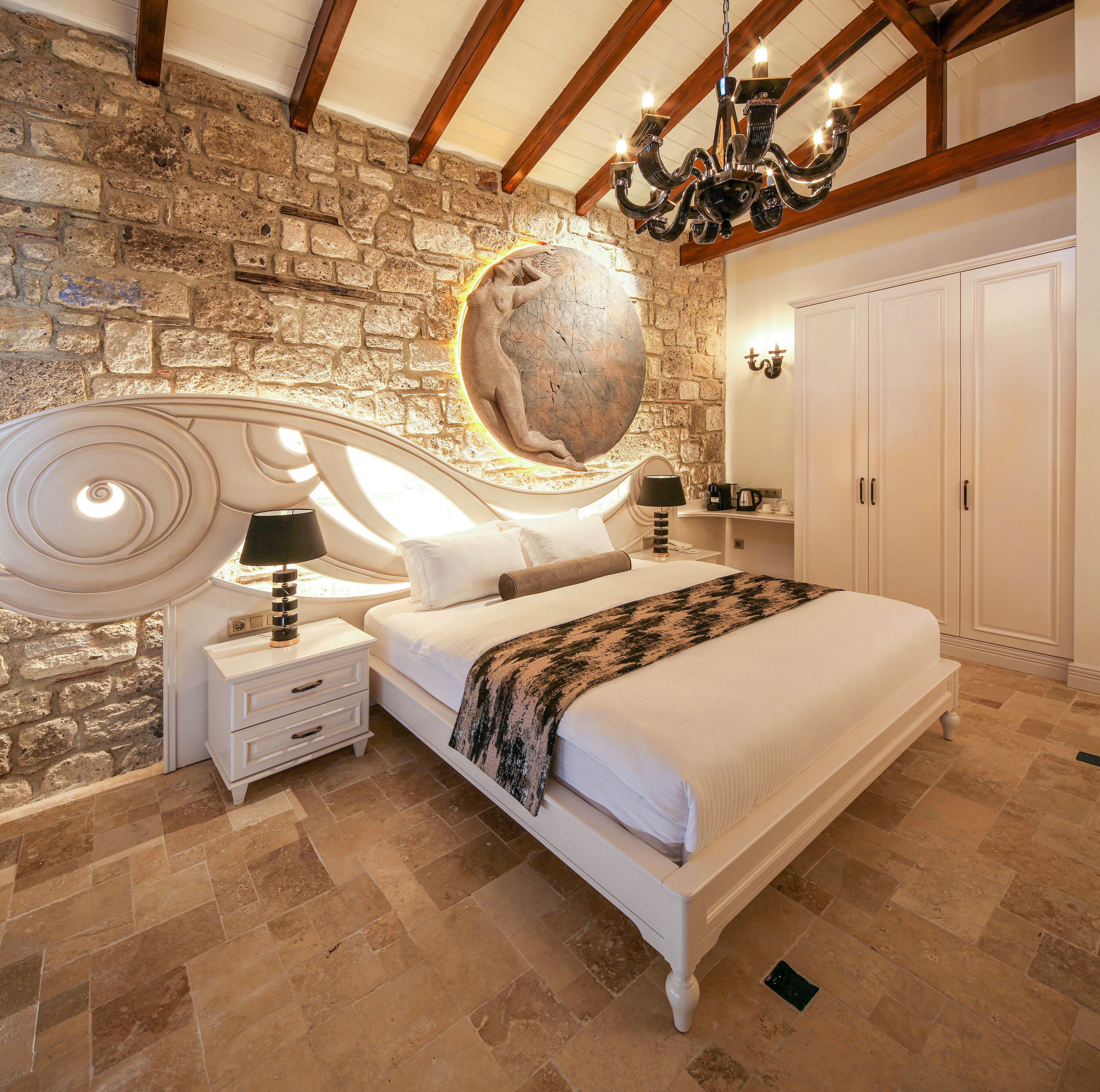 A bedroom with a stone wall, a large bed, two bedside tables with lamps, a chandelier, and a wardrobe.