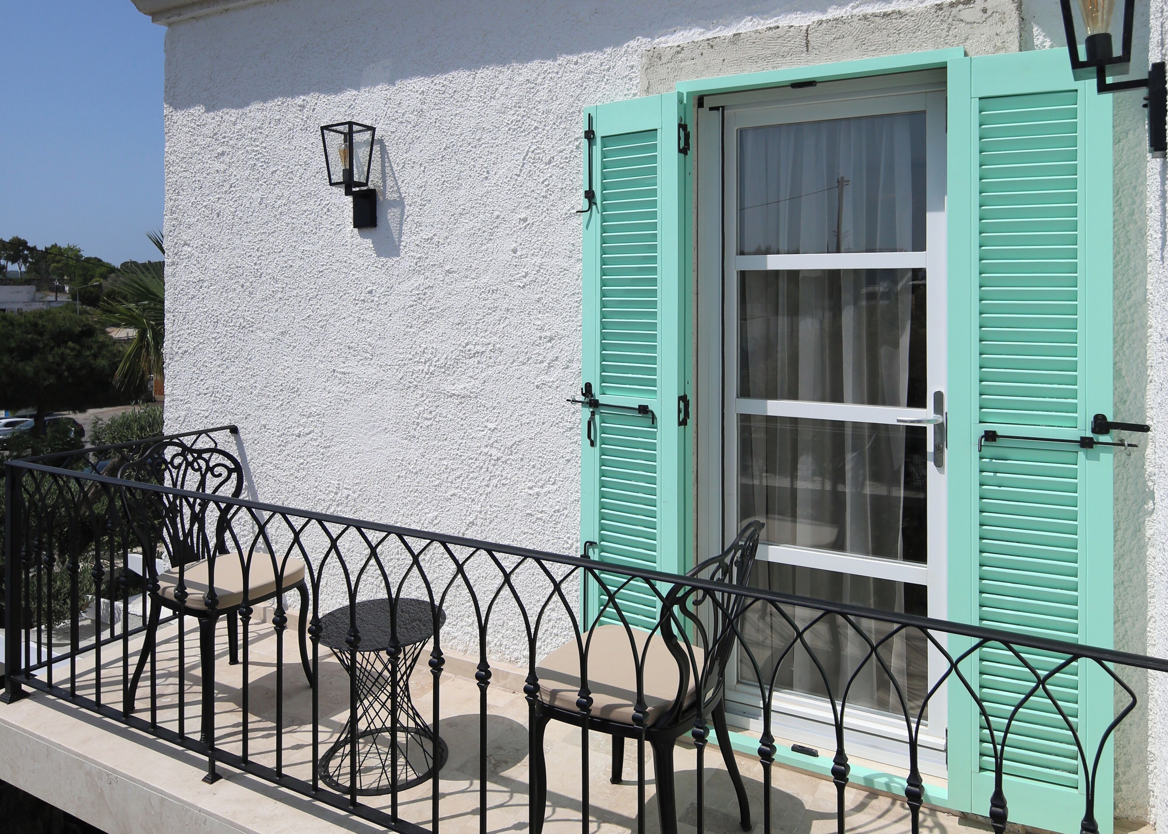 A balcony with a table and chairs, a white wall, and a window with green shutters.