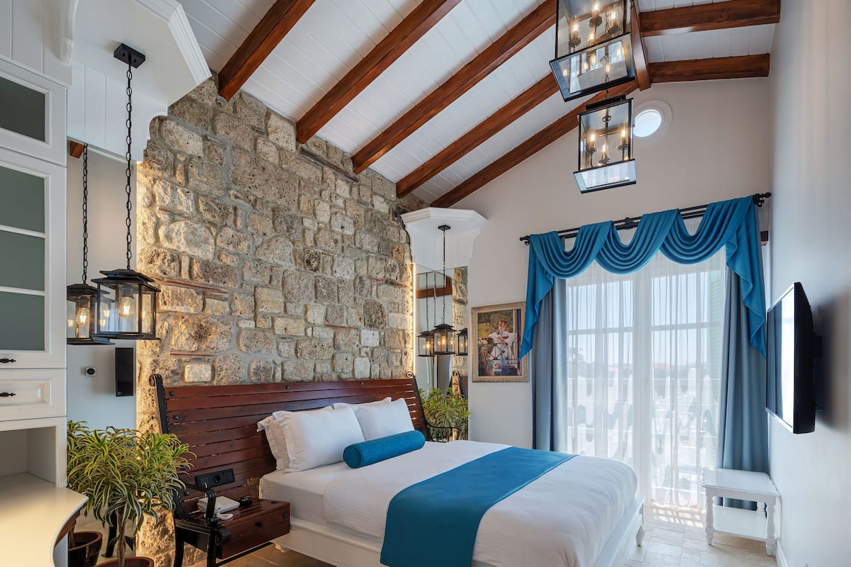 A bedroom with a stone wall, wooden beams, a bed with a blue throw, and a hanging light fixture.